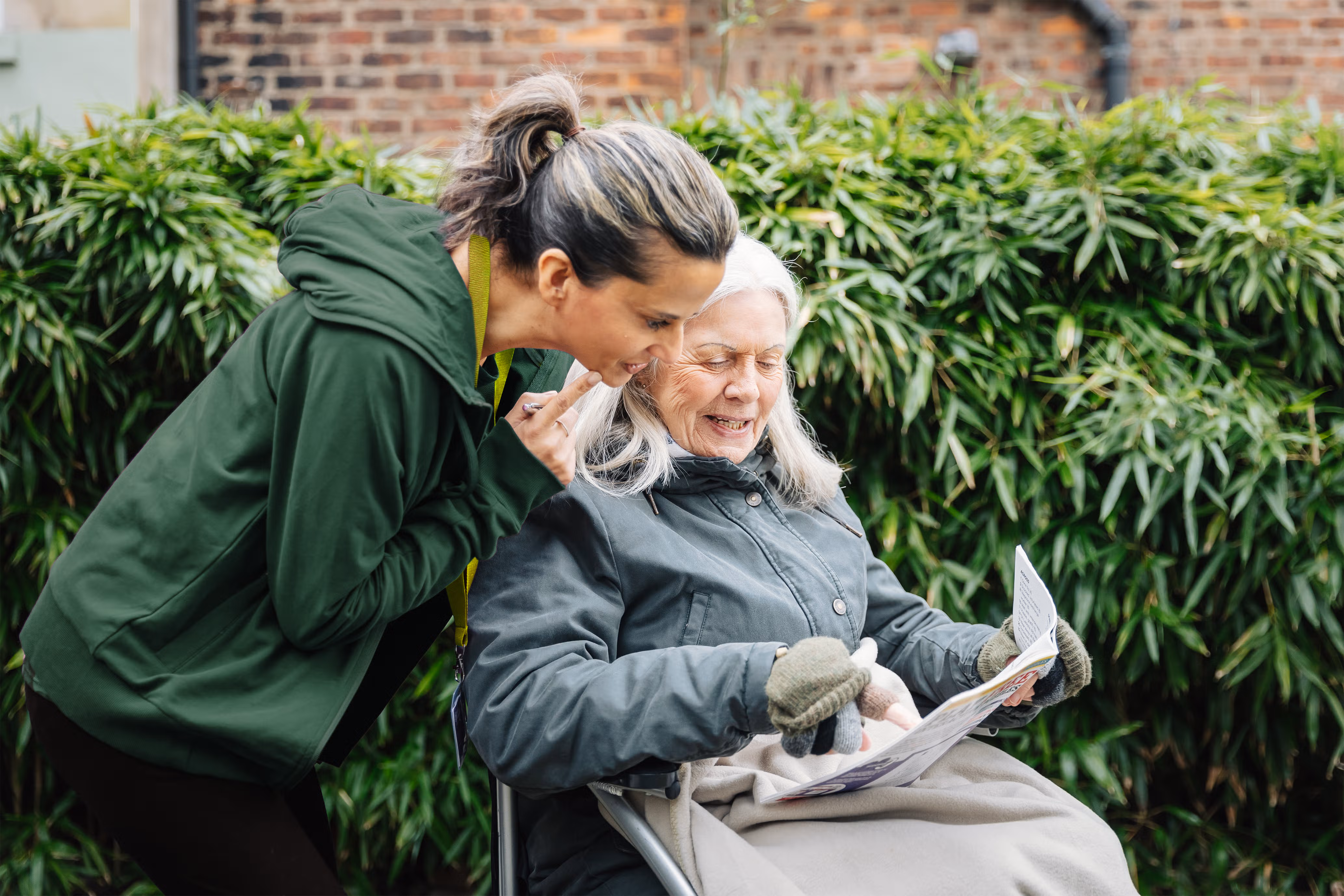 Young woman smiles whilst talking to an elderly woman reading a newspaper outdoors in a garden. - Home Instead