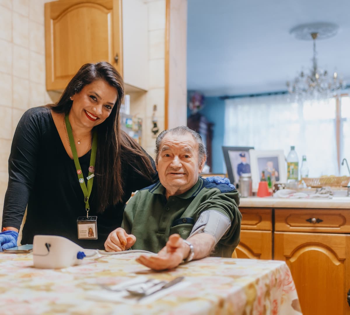 A smiling carer stands beside an elderly man seated at a kitchen table during a blood pressure check. - Home Instead