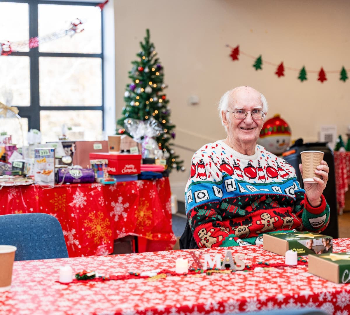 An older male adult with white hair and wearing eyeglasses and christmas sweater with a table and christmas tablecloth and christmas trees and banners and gifts at the back