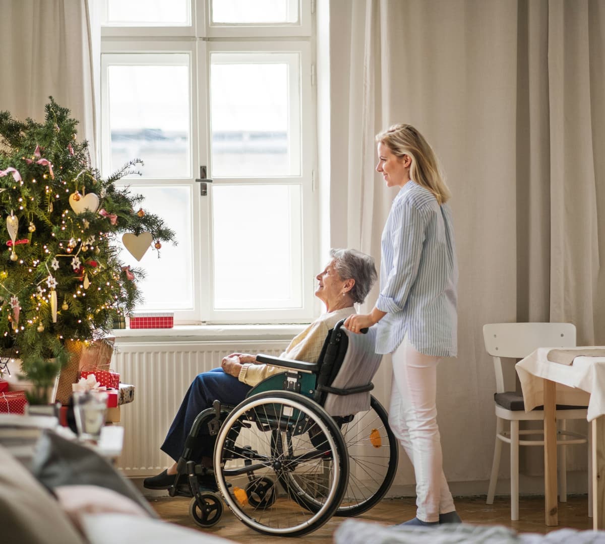 An older female adult with short grey hair sitting on a wheelchair while looking at the window and the Christmas tree inside her house with her younger female carer wearing long sleeves and pants