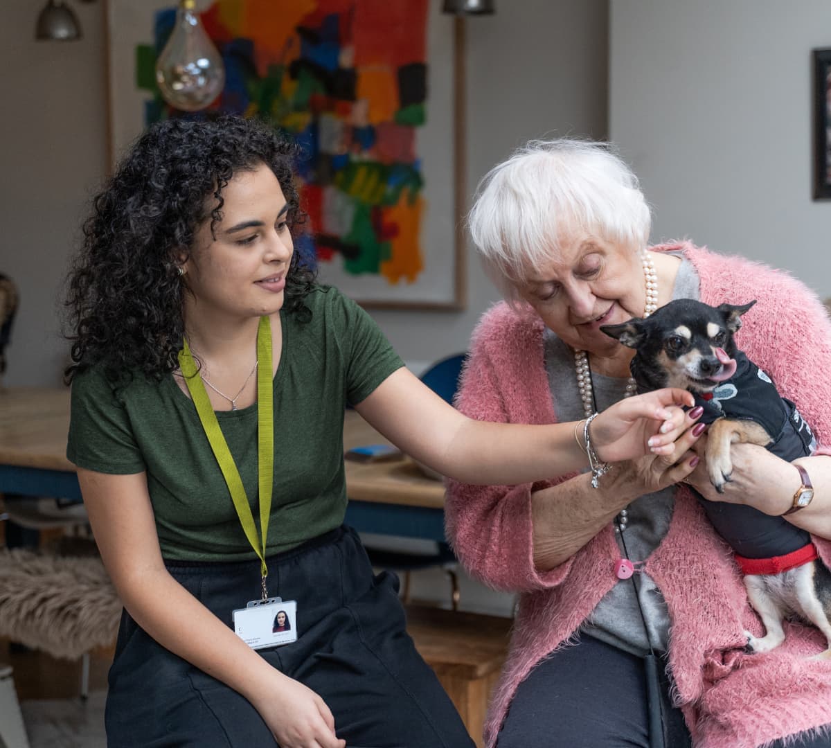 A young woman and an elderly woman smile whilst holding a small dog in a cosy, brightly decorated room. - Home Instead