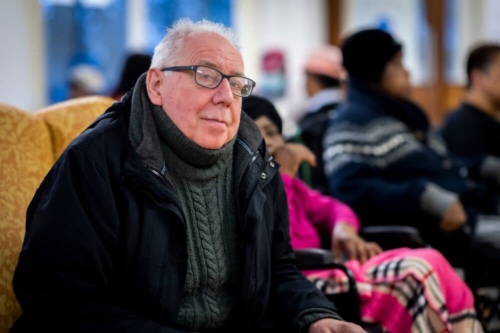Older man in spectacles and winter clothes sits indoors, with others in the background. - Home Instead