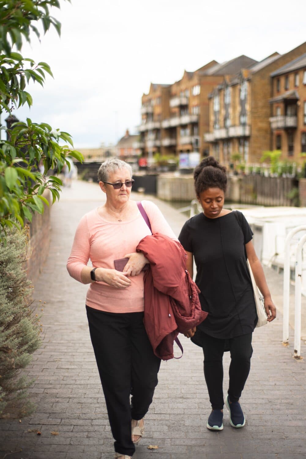 Two women walk together on a riverside path, passing flats on a cloudy day. - Home Instead