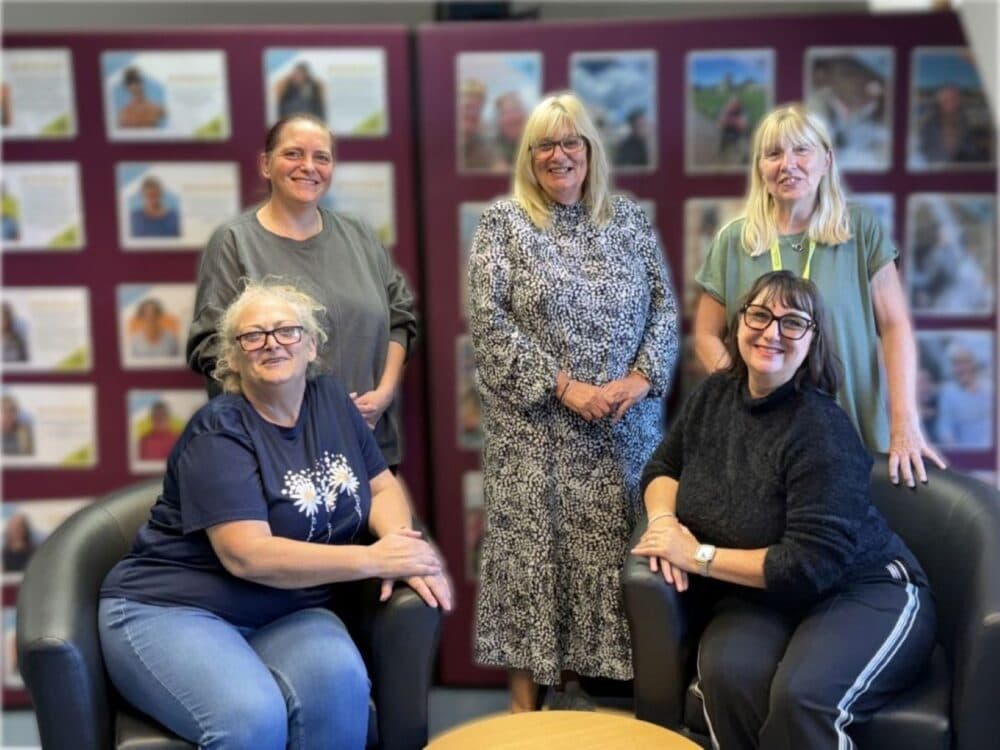 Five smiling women pose indoors in front of a wall covered with photos and posters. - Home Instead
