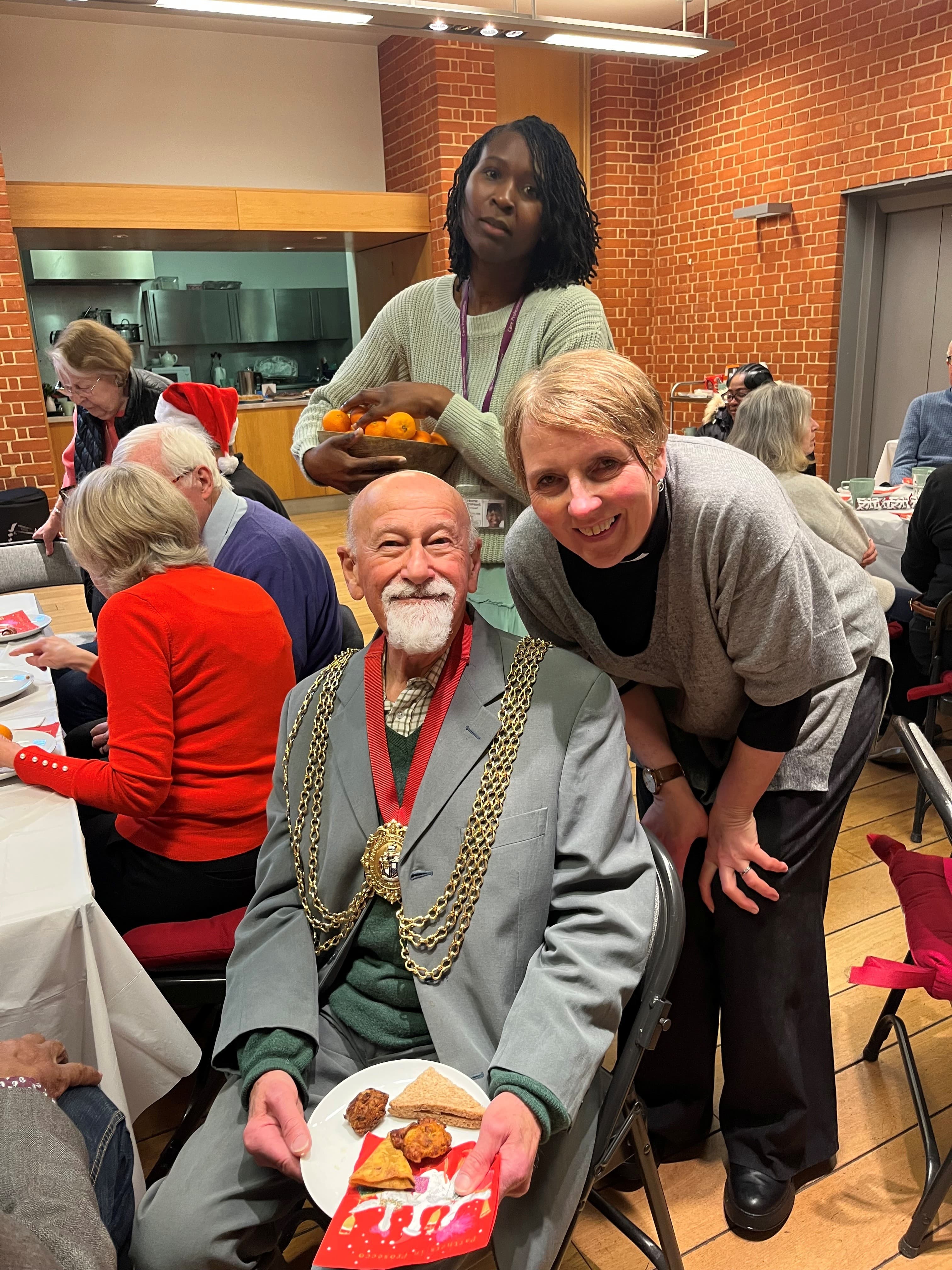 A smiling man in formal attire sits with a plate of food at a festive gathering, surrounded by others. - Home Instead