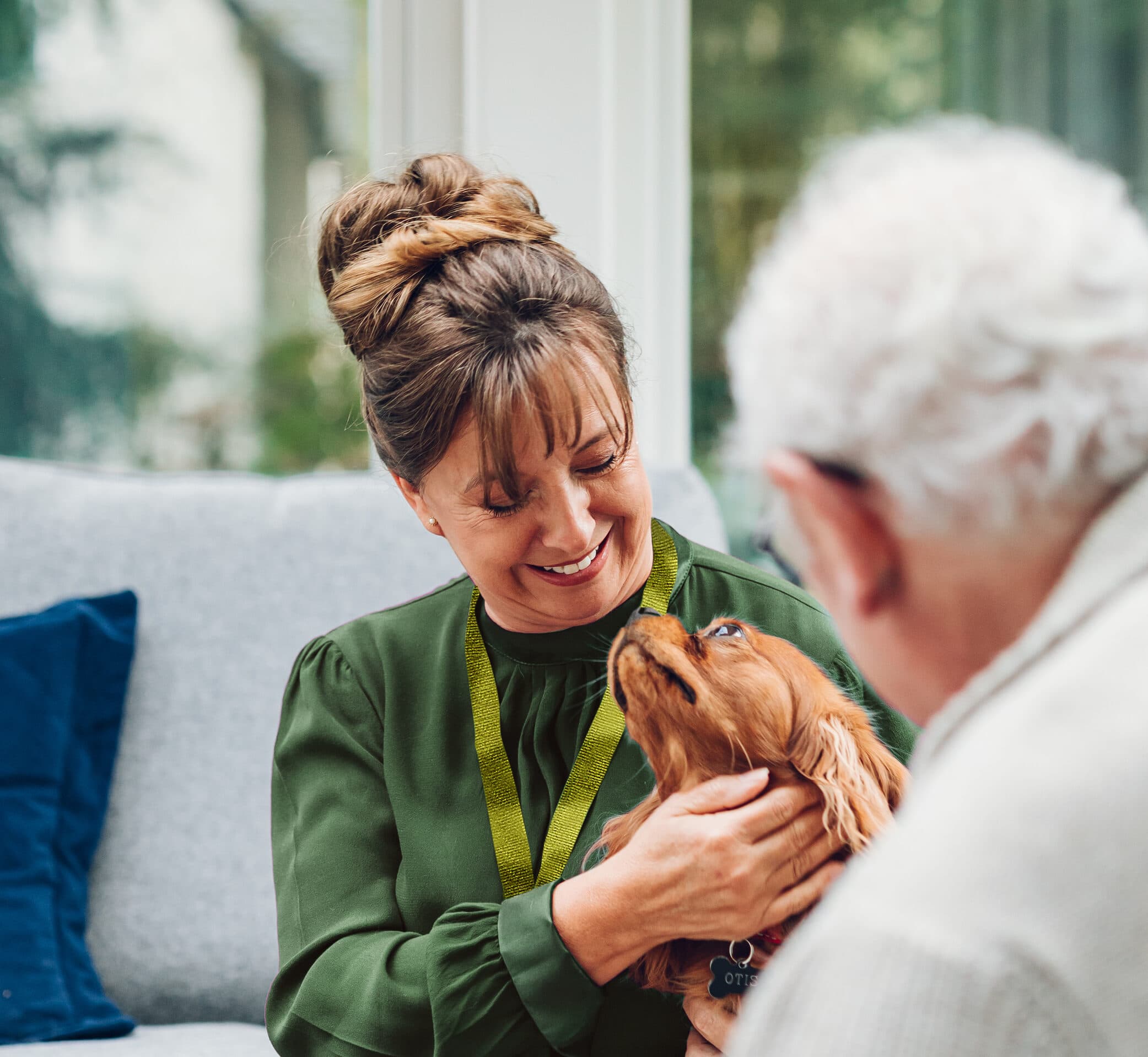 A woman smiles whilst stroking a dog, sitting beside an older man indoors. - Home Instead