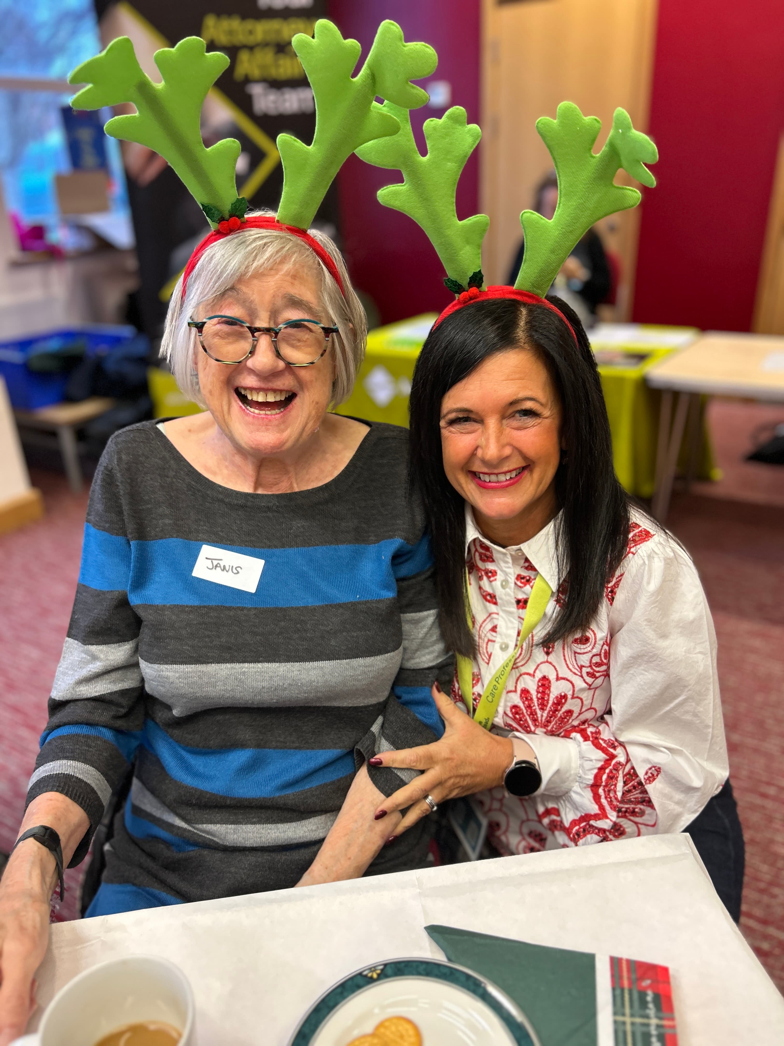 Two women smiling at a table, both wearing green reindeer antler headbands at a festive indoor event.