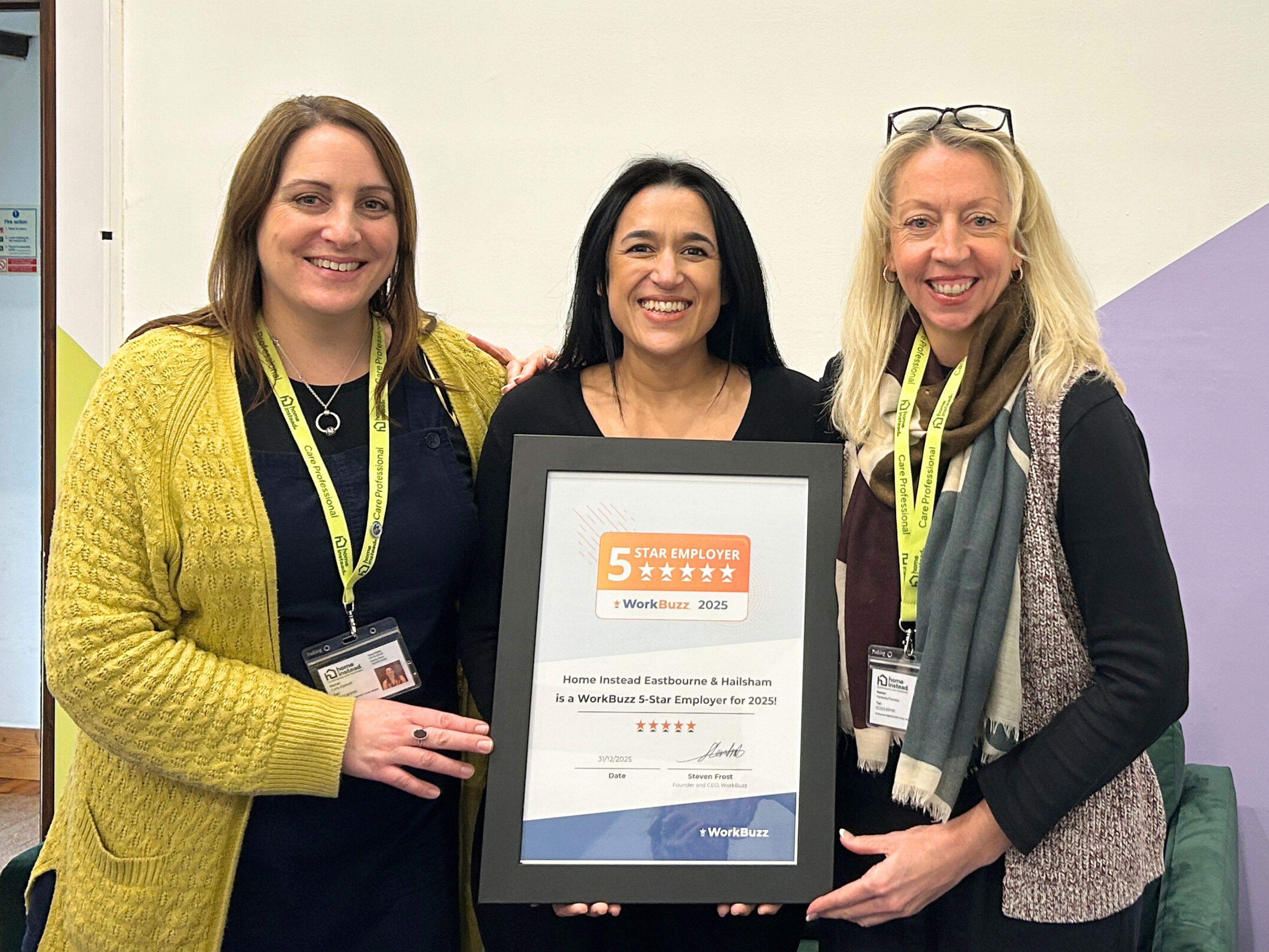 Three smiling colleagues stand indoors holding a framed WorkBuzz 5-Star Employer 2025 certificate for Home Instead Eastbourne & Hailsham.