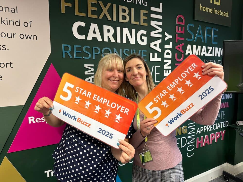 Two women smiling and holding "5 Star Employer 2025" signs in front of a colourful, positive-word wall. - Home Instead