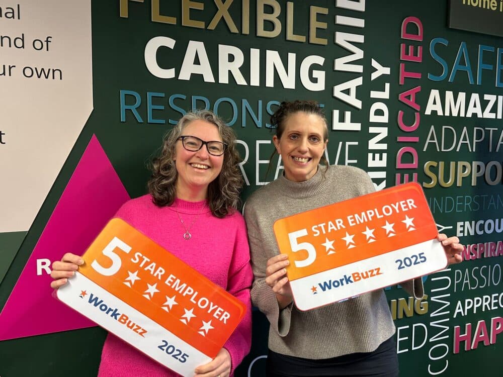 Two women smiling and holding "5 Star Employer" signs from WorkBuzz 2025 in front of a colourful wall. - Home Instead