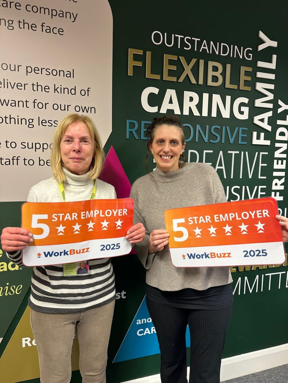 Two women smiling and holding "5 Star Employer WorkBuzz 2025" signs, standing by a wall with positive words. - Home Instead