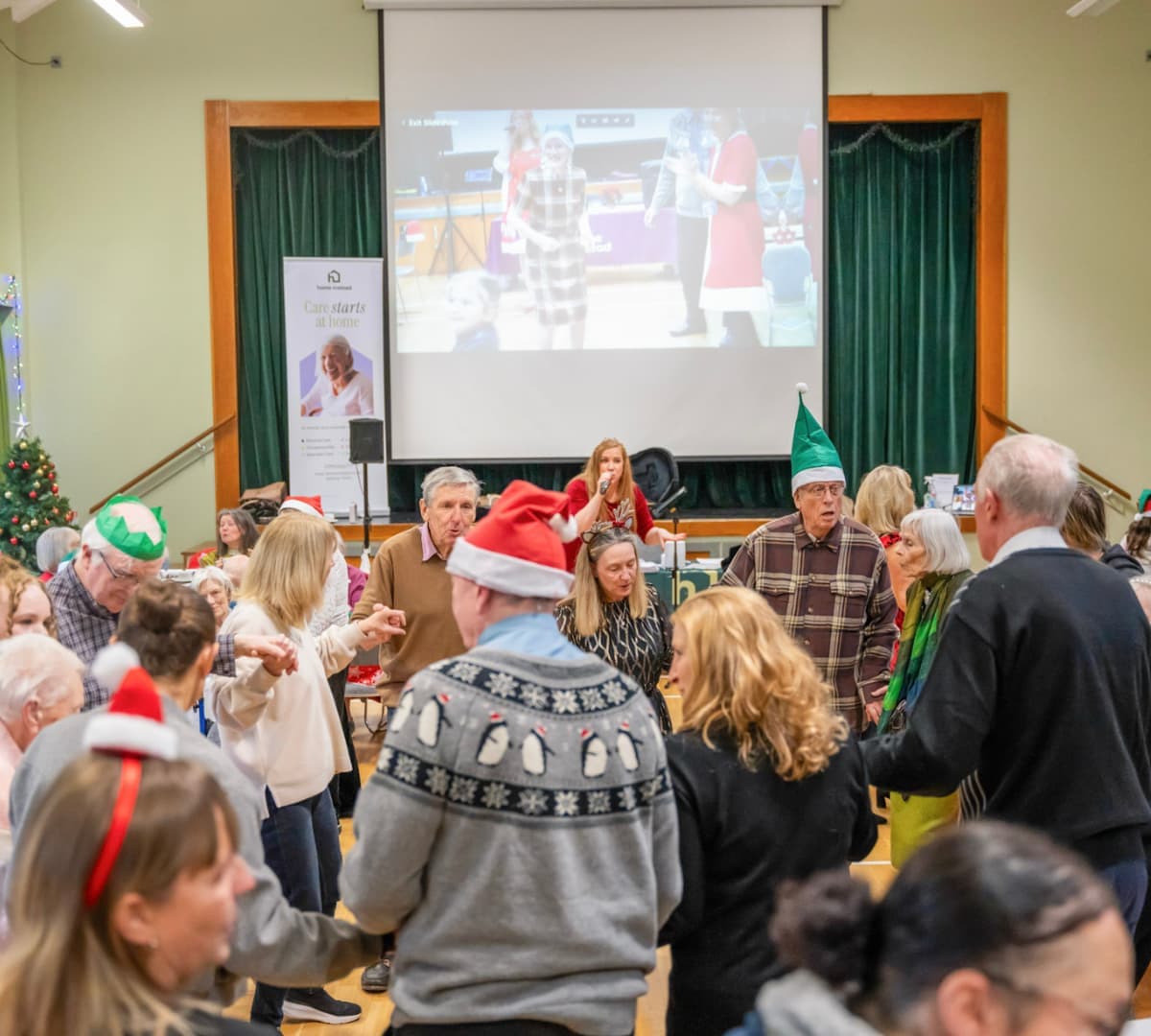 People wearing festive hats dancing in a decorated hall with a Christmas tree and a large screen in the background. - Home Instead