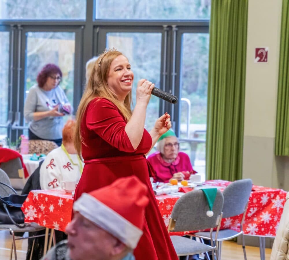 Woman in a red dress sings with a microphone at a festive gathering with holiday decorations and guests. - Home Instead