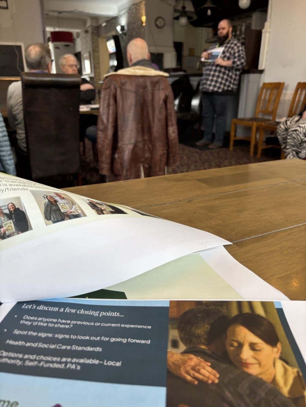A man speaks to a seated group in a cosy room; leaflets and papers are on a table in the foreground. - Home Instead