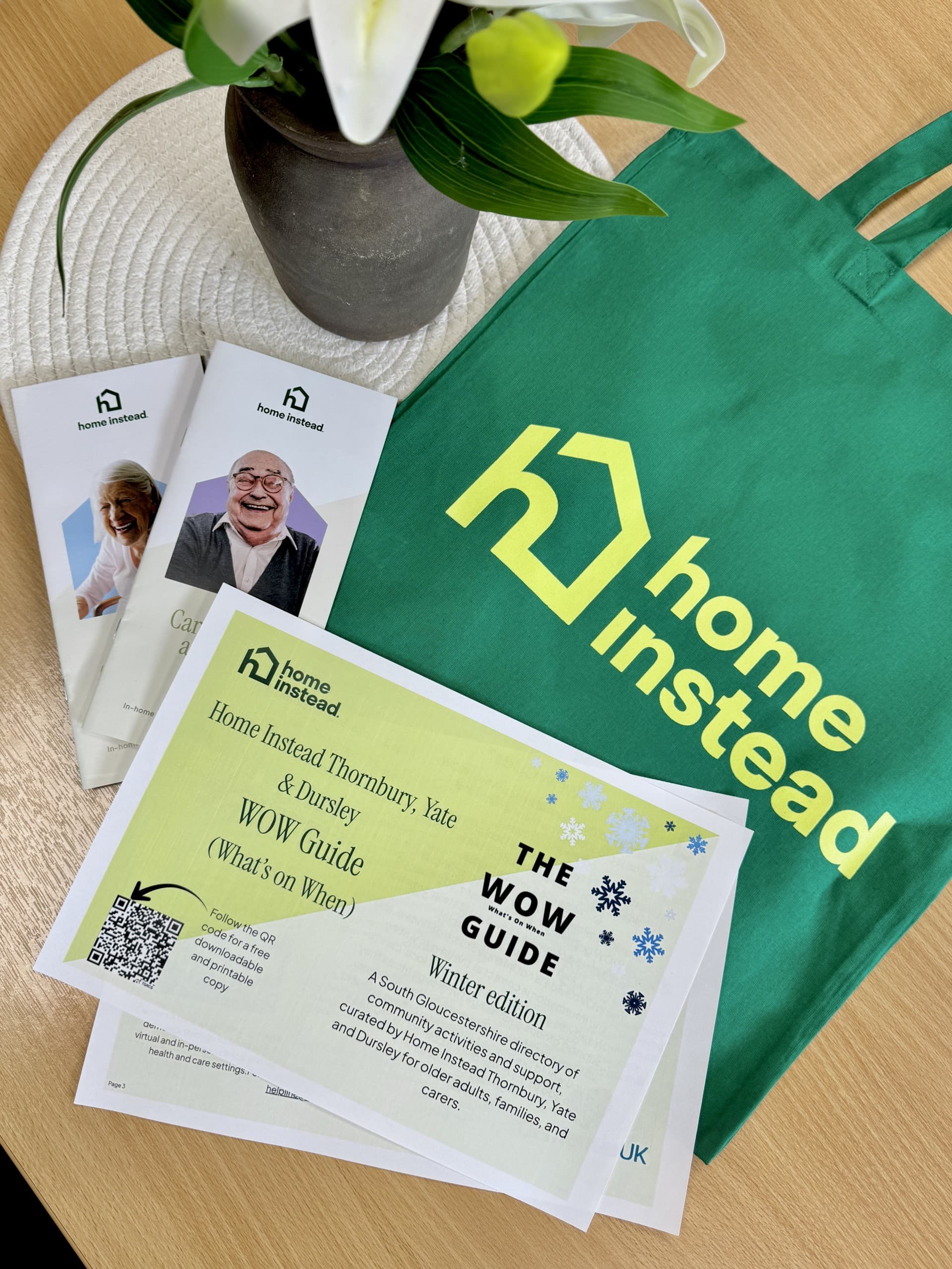 A green Home Instead tote bag, brochures, and a WOW Guide are displayed on a table with a white flower. - Home Instead