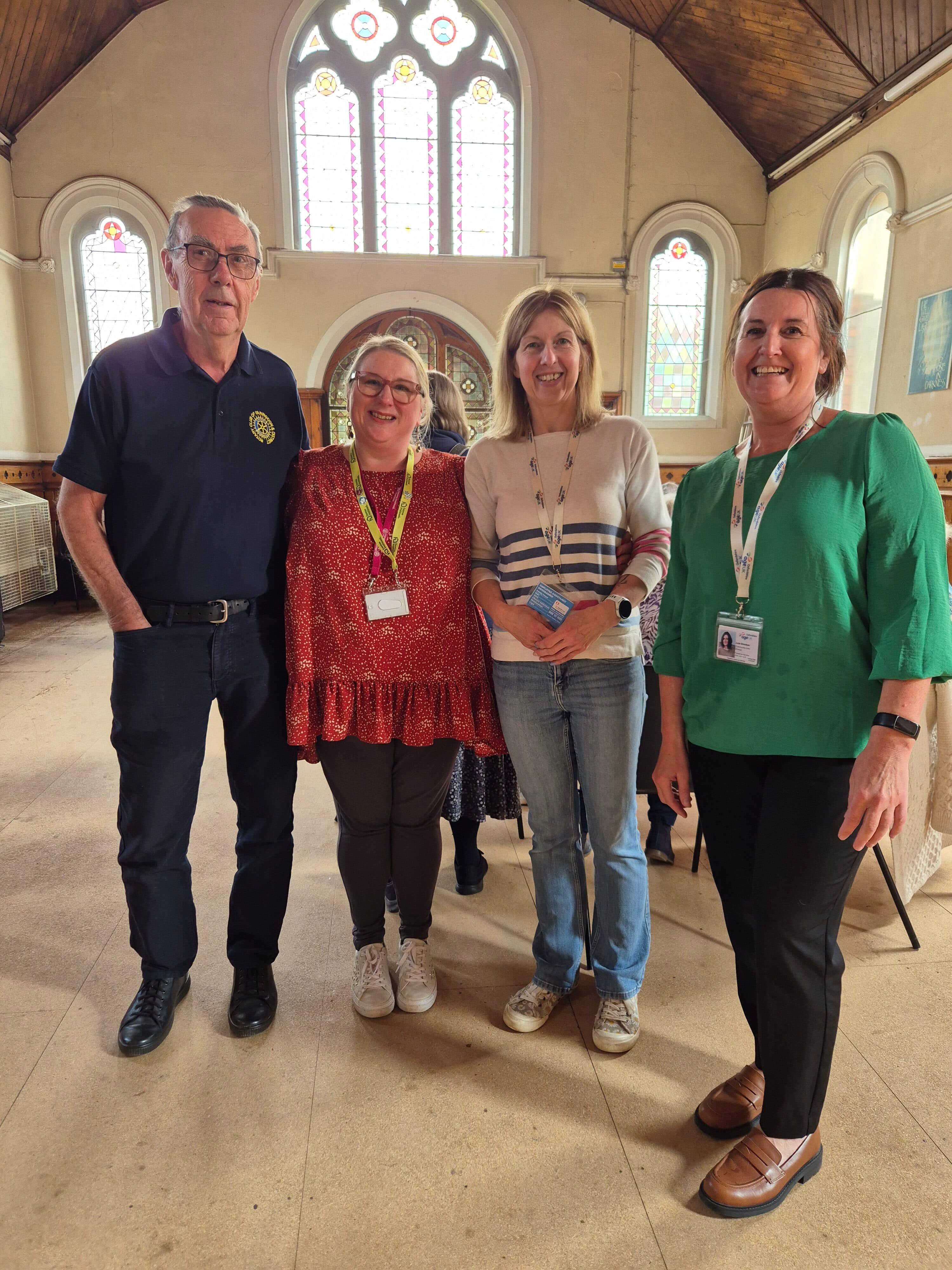 Four adults stand smiling inside a hall with arched windows and stained glass in the background. - Home Instead