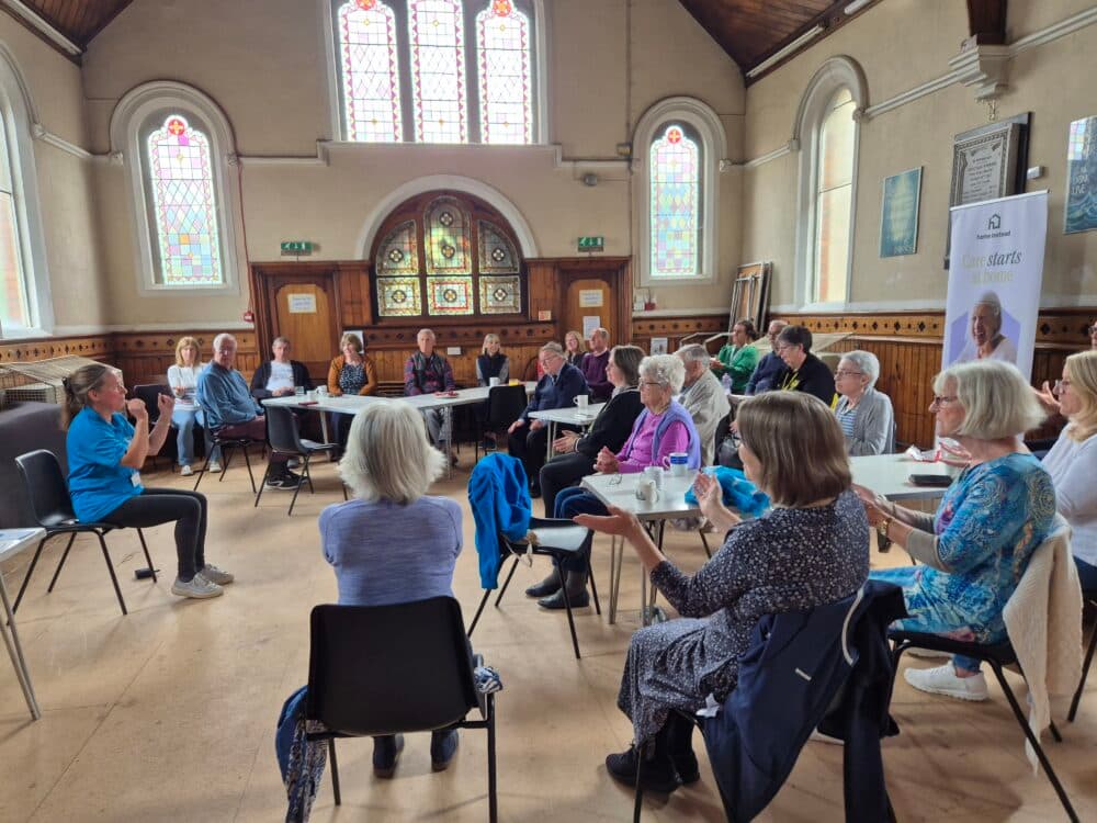 A group of people sit in a church hall, listening to a woman speaking at the front of the room. - Home Instead