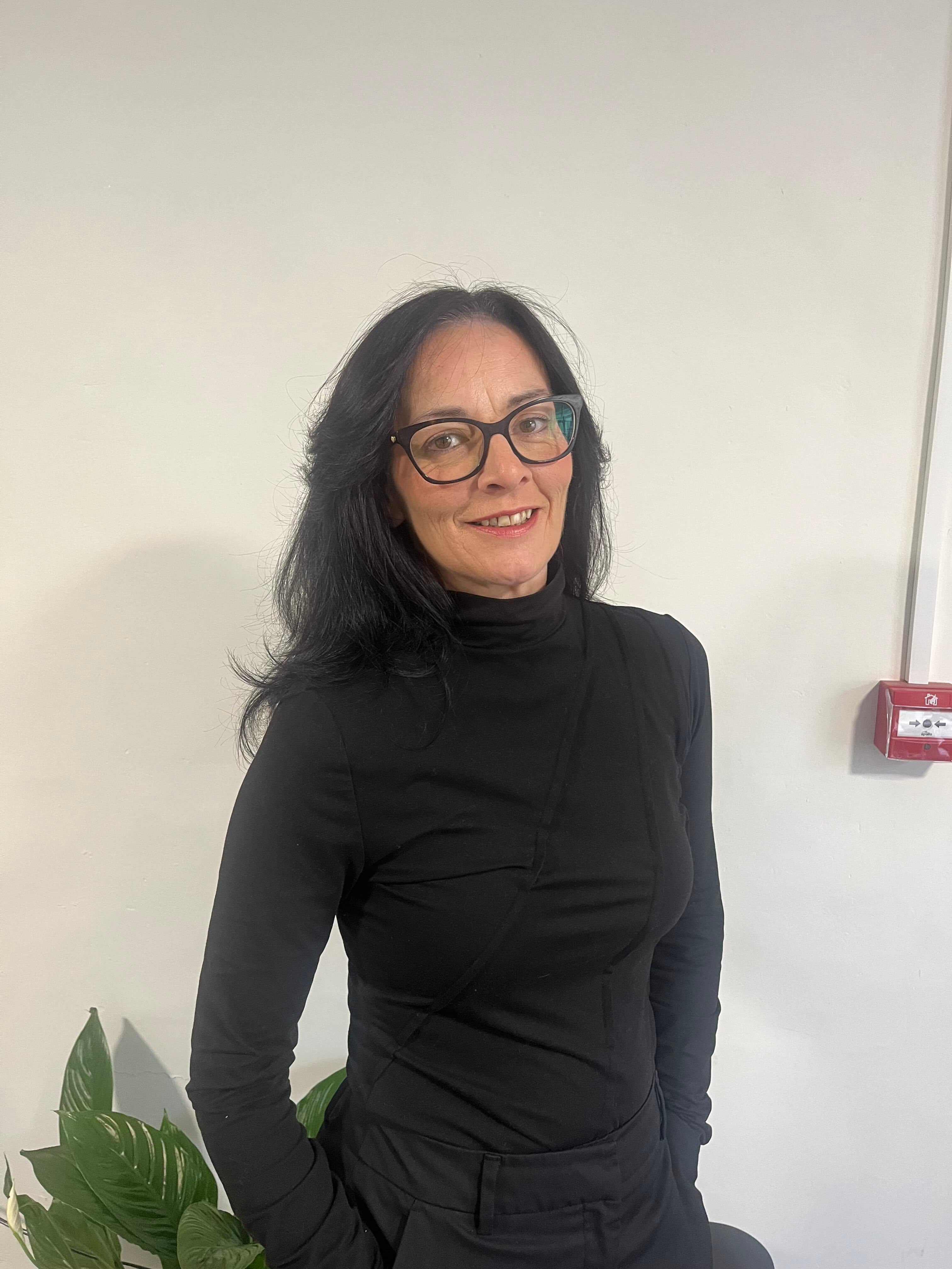 Woman with long dark hair and glasses in black clothes, standing indoors near a plant and a fire alarm on the wall. - Home Instead