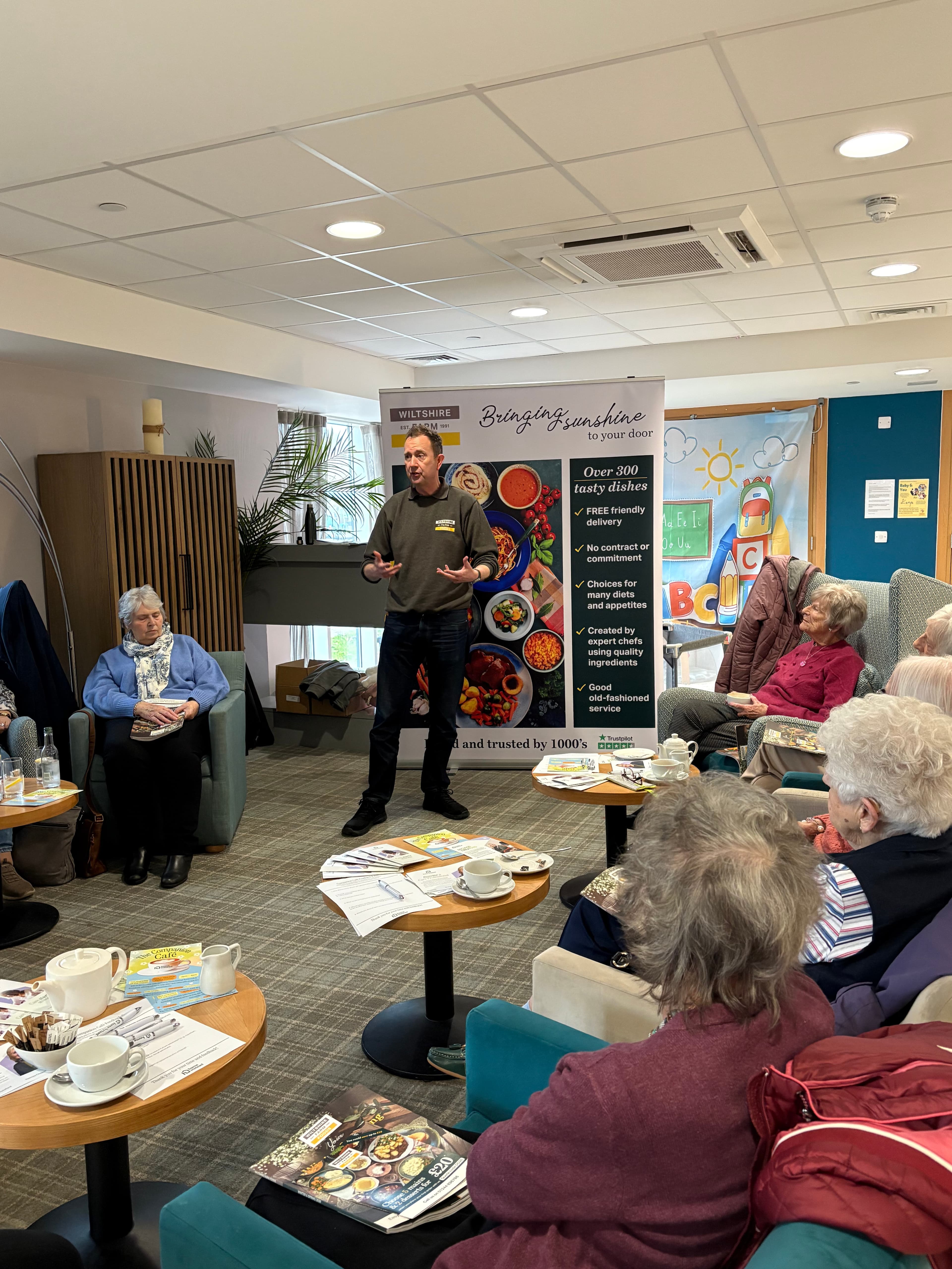 A man speaks to a group of elderly people seated round tables with tea and magazines in a cosy room. - Home Instead