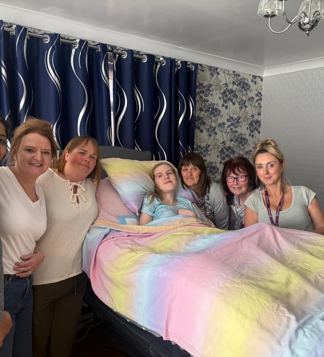 Six women gather around a young girl in bed, smiling together in a cosy, homely room with patterned curtains and walls. - Home Instead