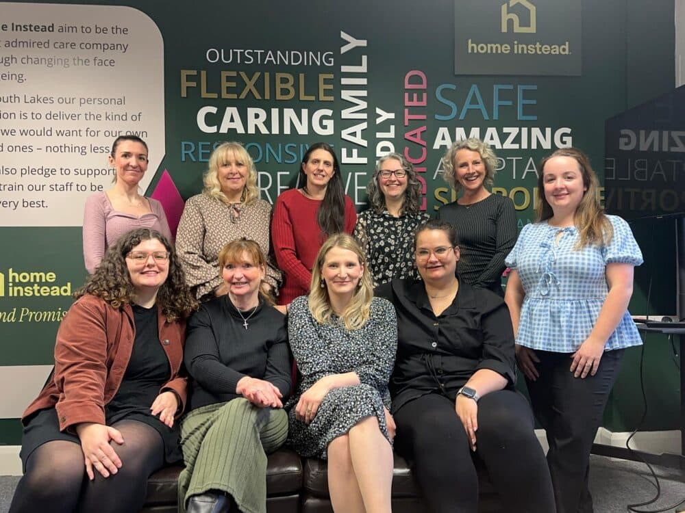 A group of eleven women smiling and posing together in an office with motivational words on the wall behind them. - Home Instead
