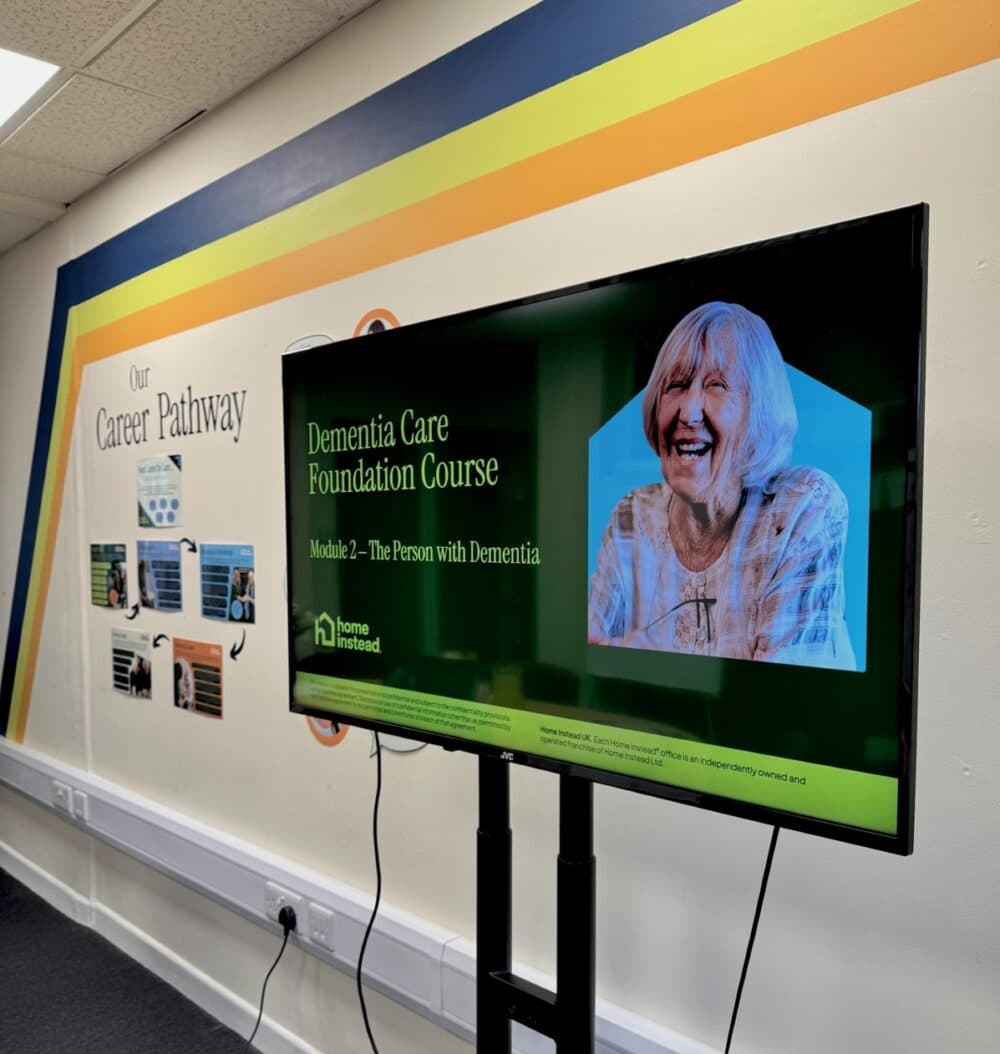 TV displays "Dementia Care Foundation Course" beside a wall with "Our Career Pathway" and photos. - Home Instead