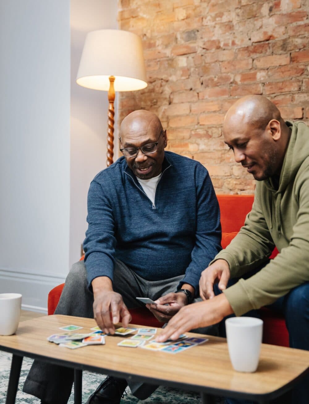 Two men sit on a red sofa playing a card game at a small table with mugs in a cosy living room. - Home Instead