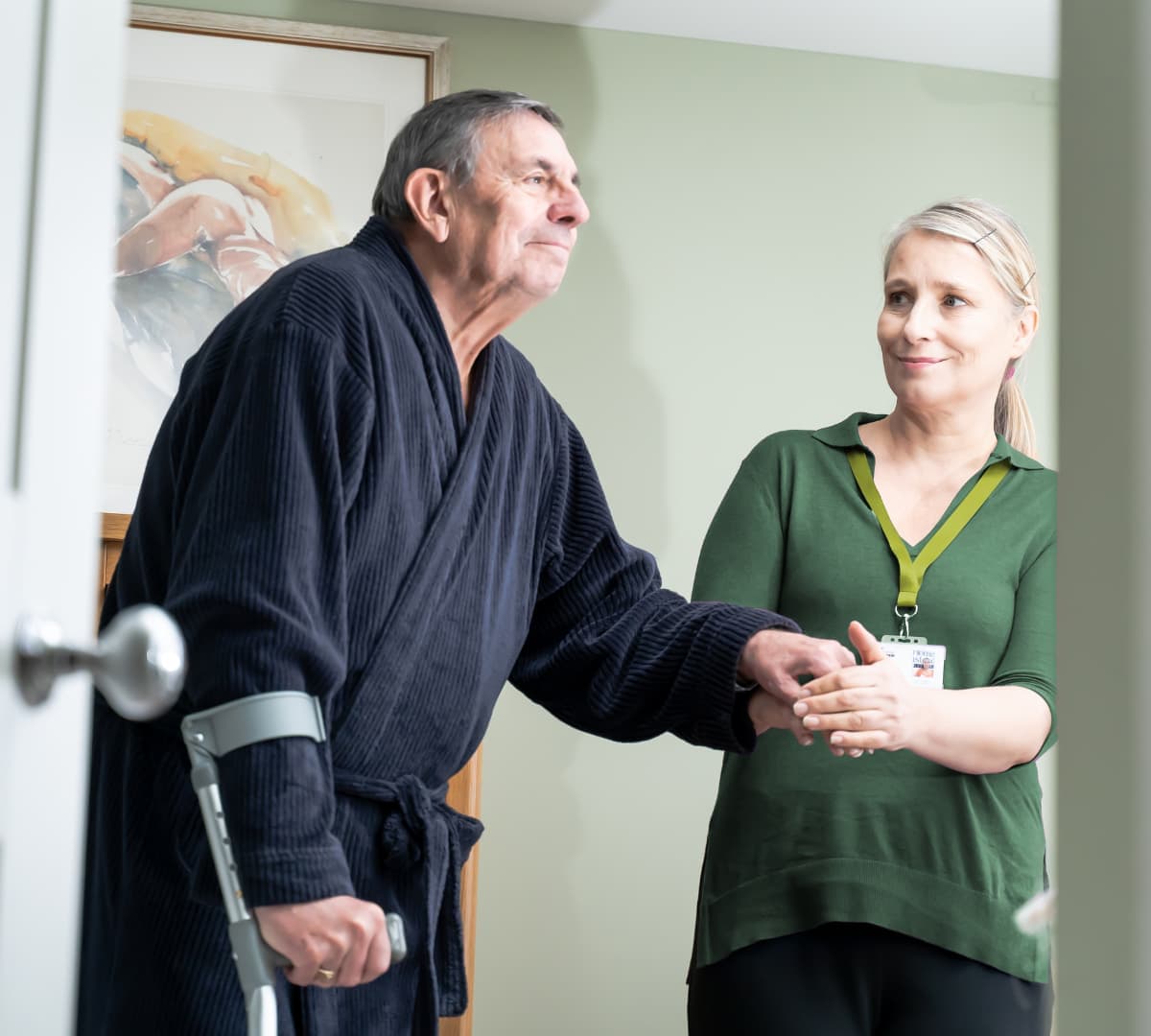 An older male adult walking while using a crutch and being helped by his younger female carer