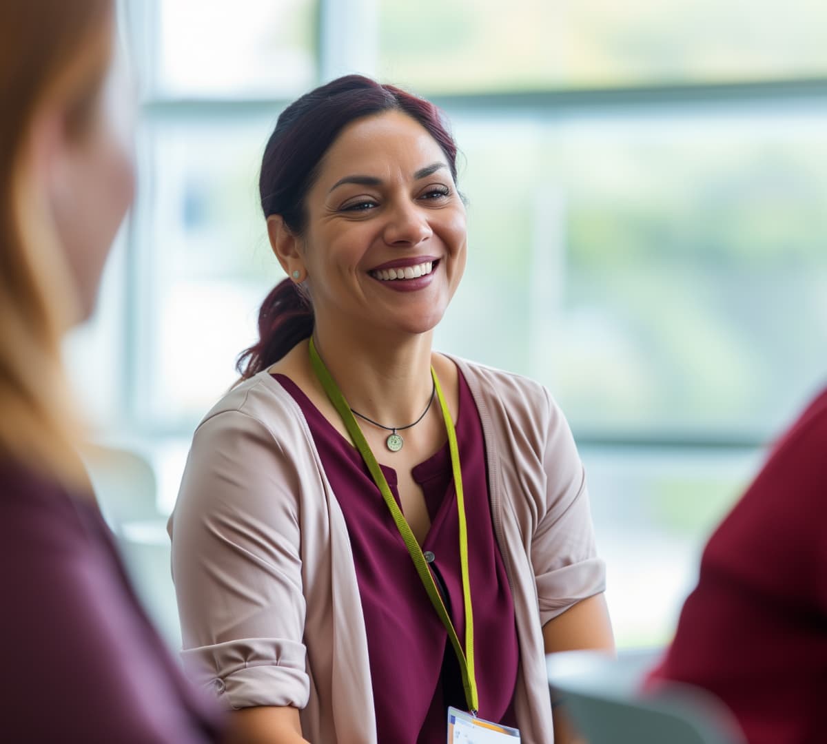 Smiling woman with dark hair sits indoors, wearing a lanyard and talking with others in a bright room. - Home Instead