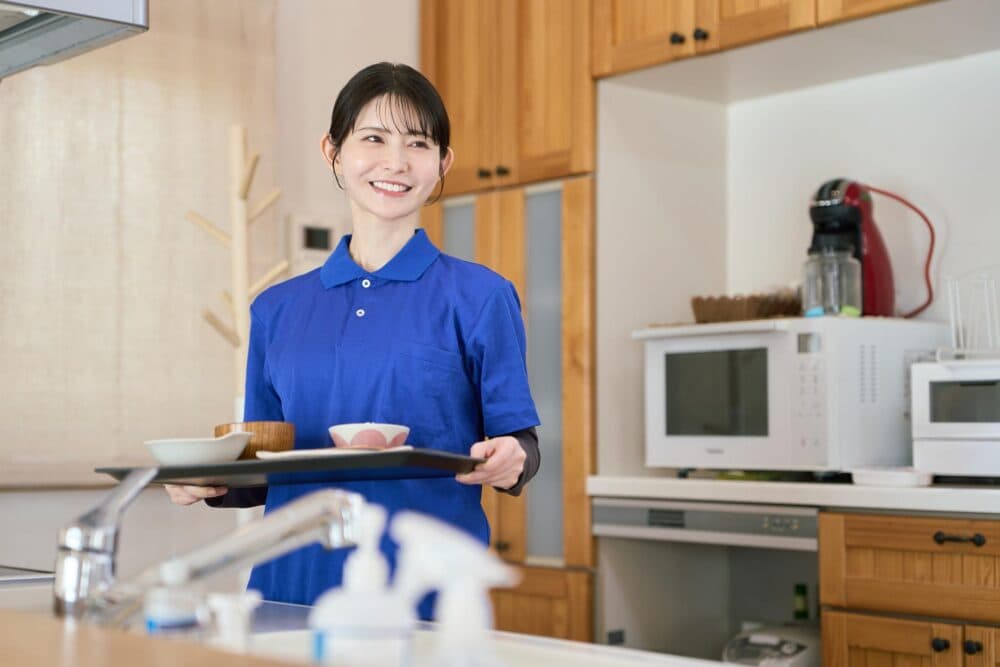 A smiling woman in a blue shirt carries a tray with food in a kitchen with wooden cupboards and appliances. - Home Instead