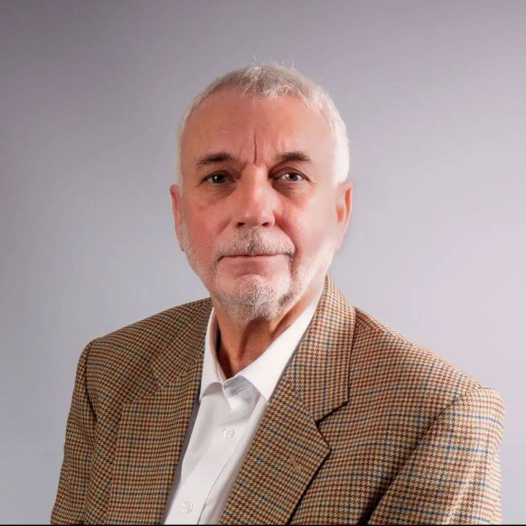 Older man with short white hair and beard, wearing a brown checked blazer and white shirt, against a grey background. - Home Instead
