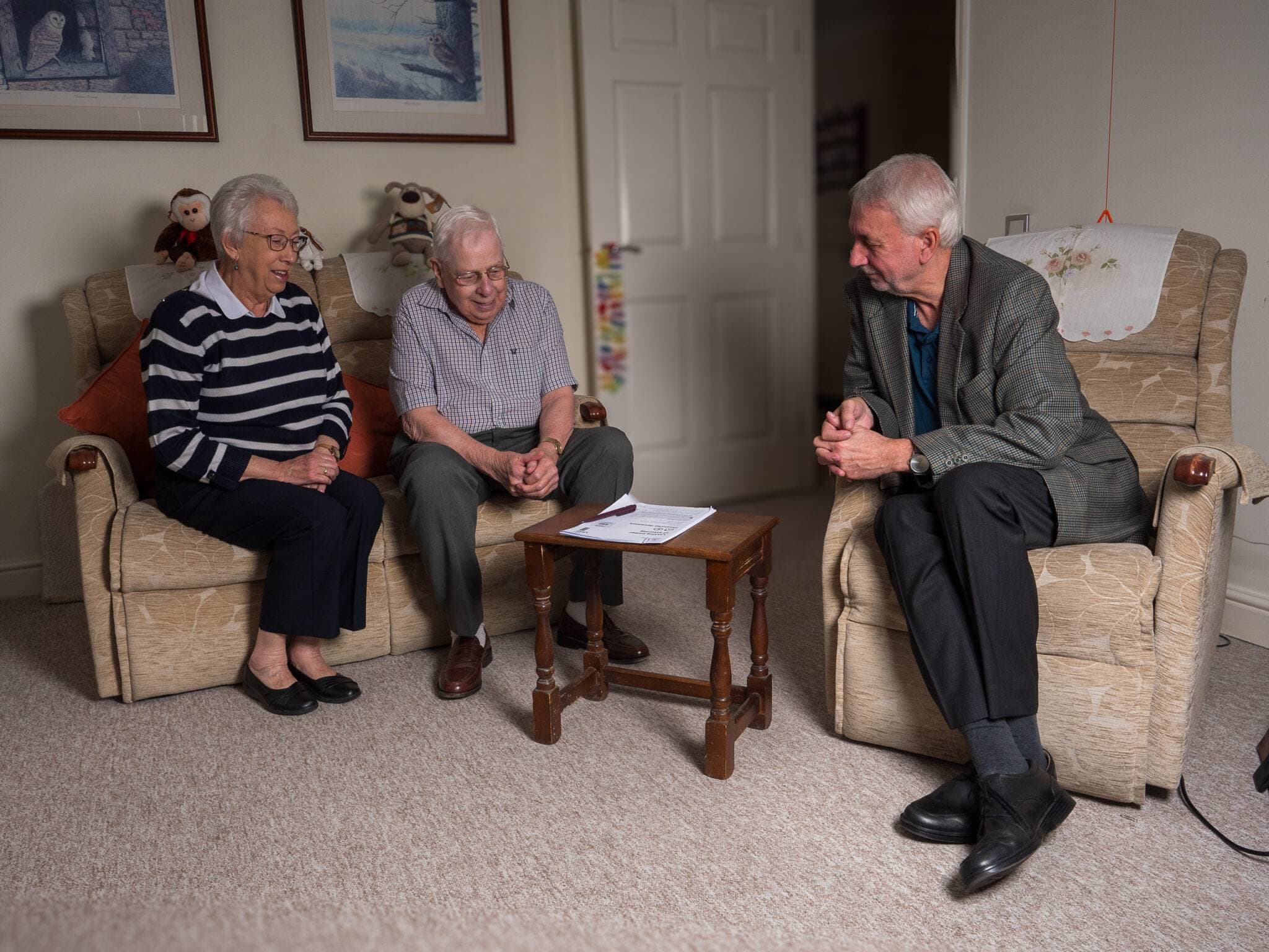 Three elderly people sit talking in a living room, with a document on a small table between them. - Home Instead
