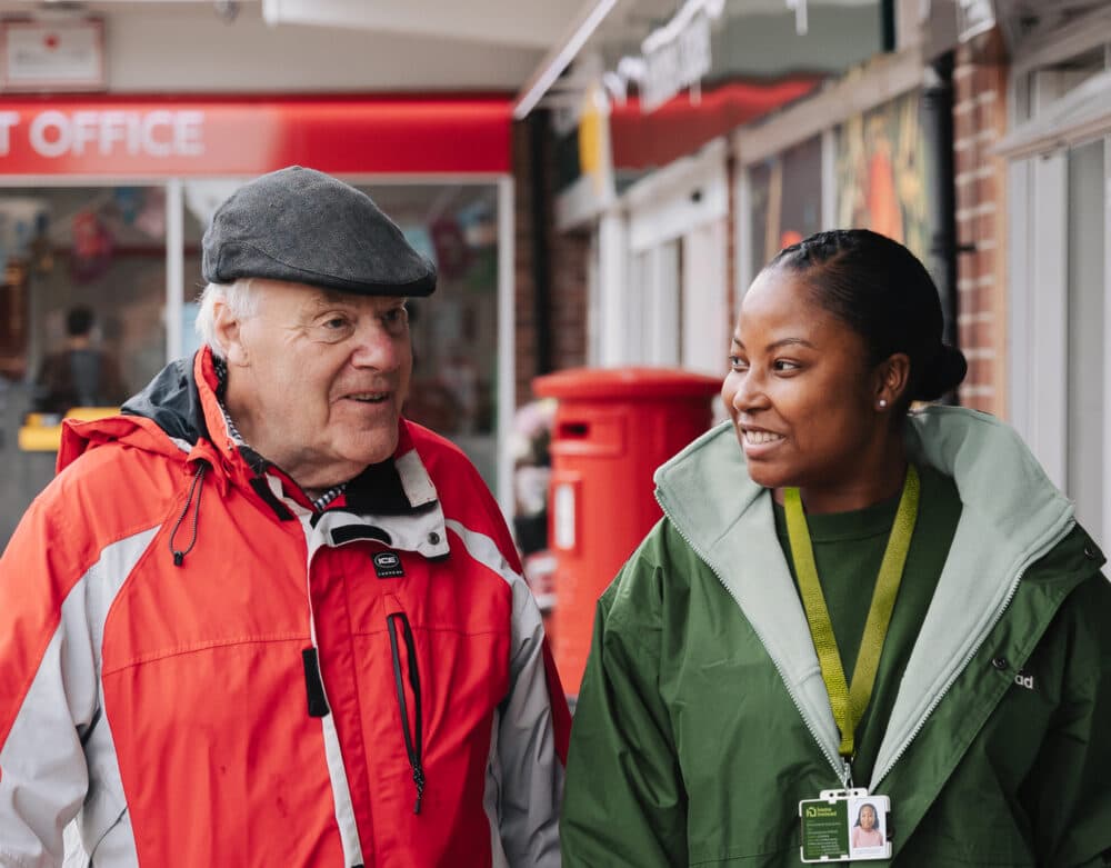 An elderly man and a young woman in jackets smile whilst walking outside near a post office. - Home Instead