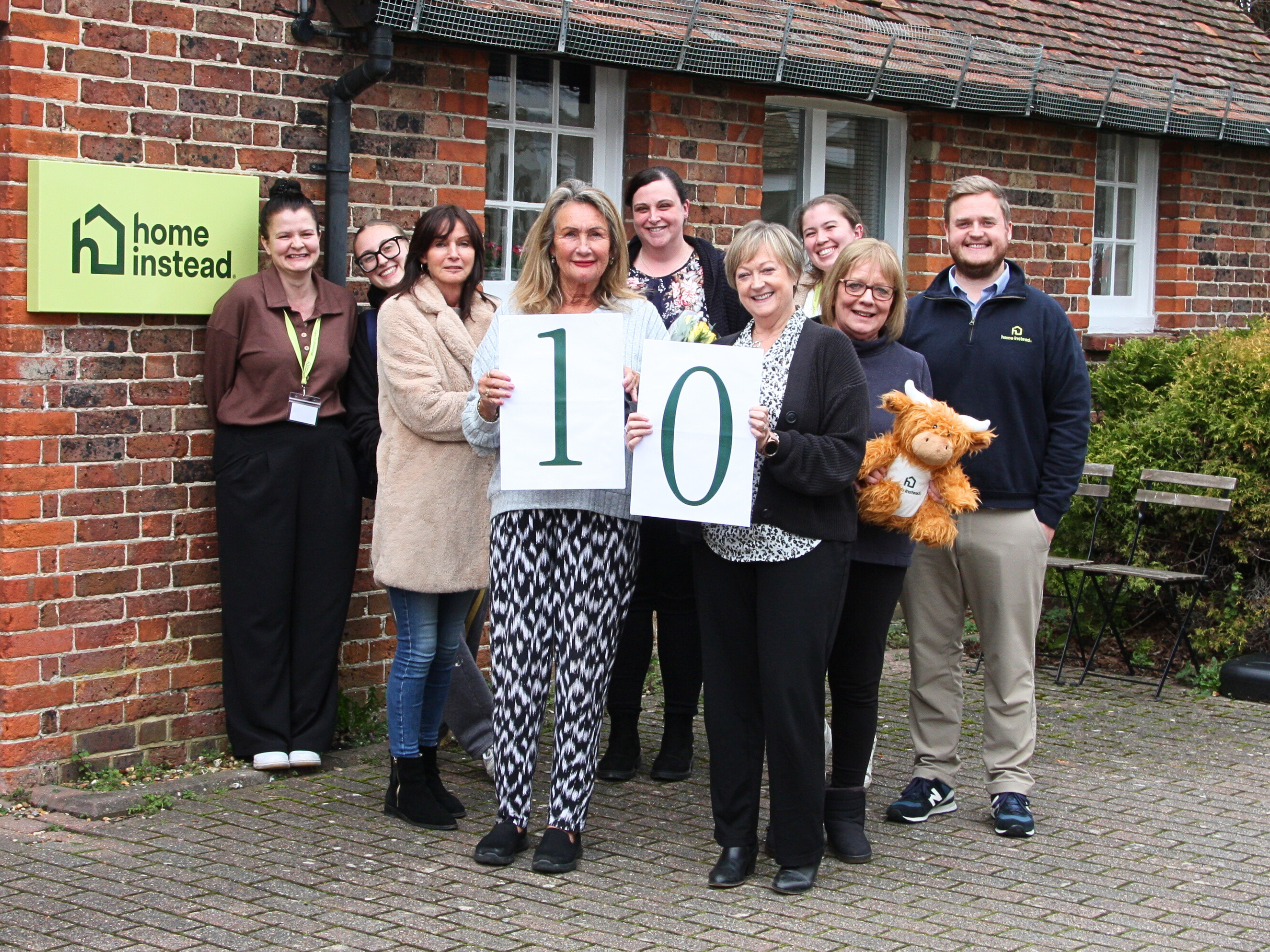 A group of smiling staff members stand outside a brick building with a “Home Instead” sign, holding large cards displaying the number 10.