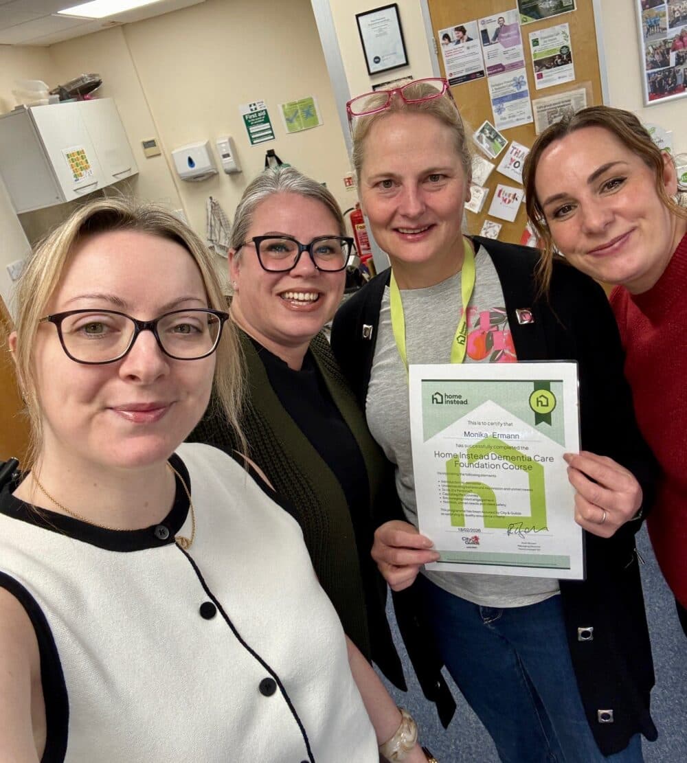 Four women smiling indoors; one holds a certificate and they pose together in an office or classroom setting. - Home Instead