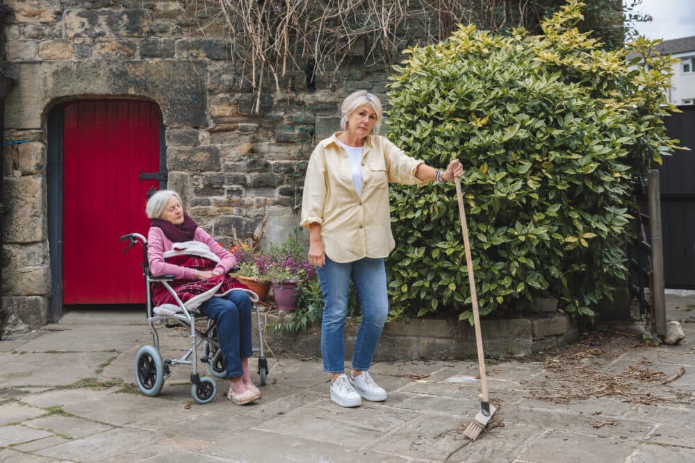 Elderly woman in a wheelchair and another woman standing with a broom outside near a stone wall and green bush. - Home Instead