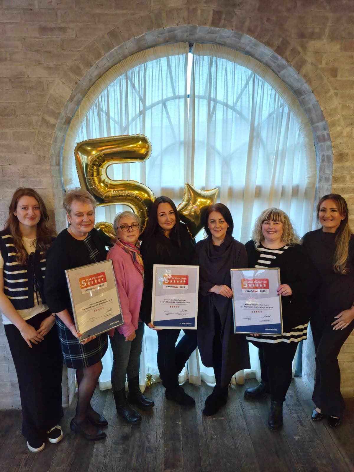 Seven women standing indoors, holding certificates, with gold "5" and star balloons in the background. - Home Instead