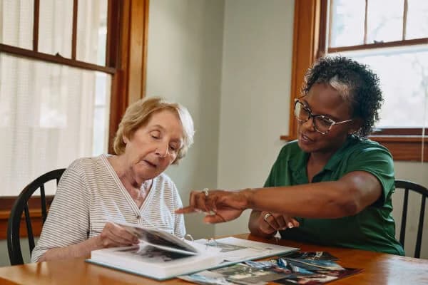 An elderly woman and a carer look at photos together at a table in a sunlit room. - Home Instead