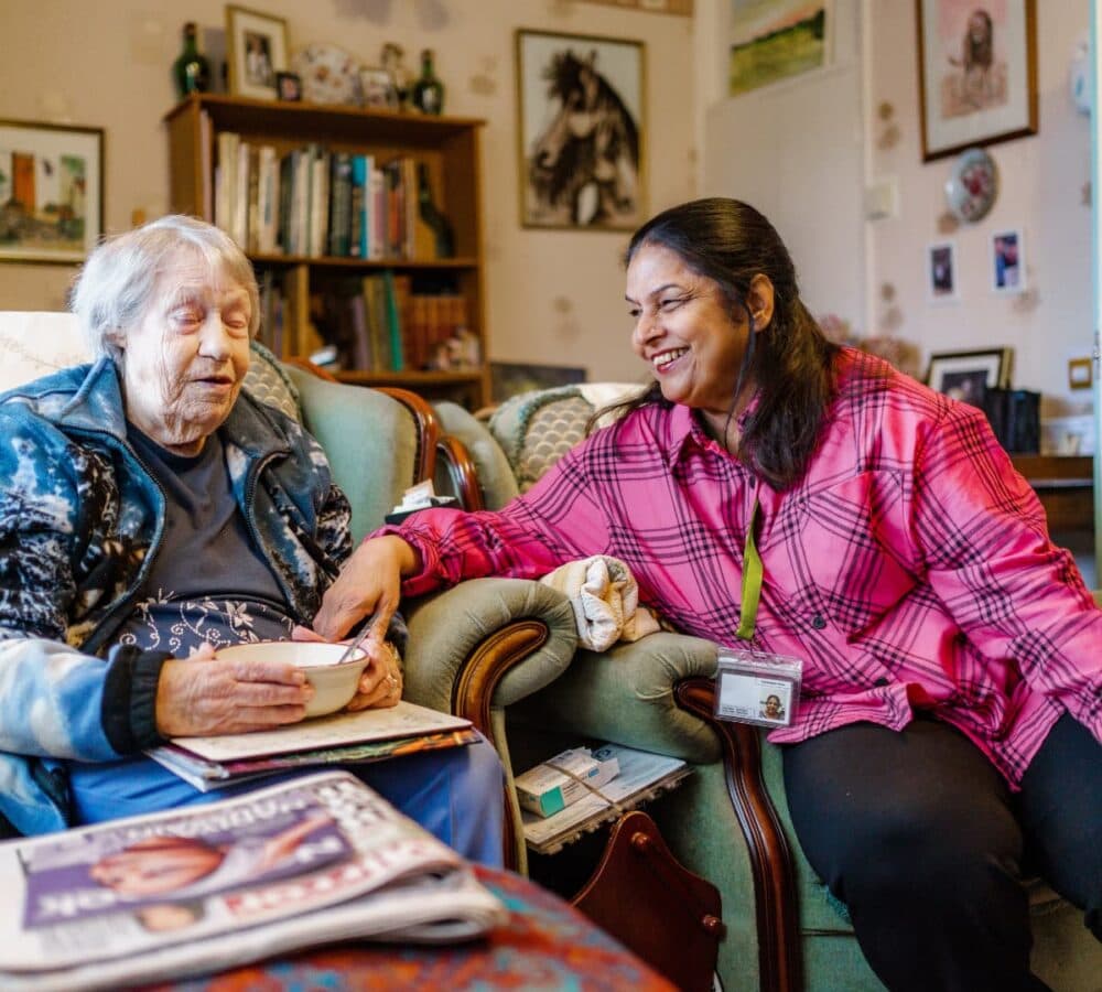 An older female adult with hrey hair eating and sitting on the couch with her younger female carer with long black hair and wearing pink inside the house