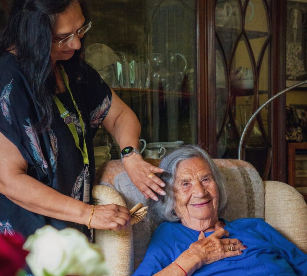 An older female adult with grey hair sitting on the couch while her hair is being combed by her carer