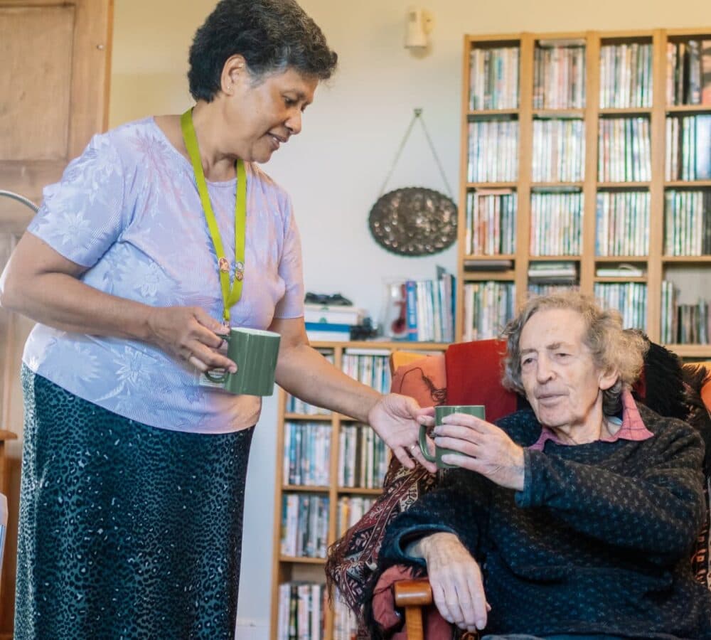 An older male adult with grey hair getting coffee from his female carer with short black hair inside the house - Home Instead