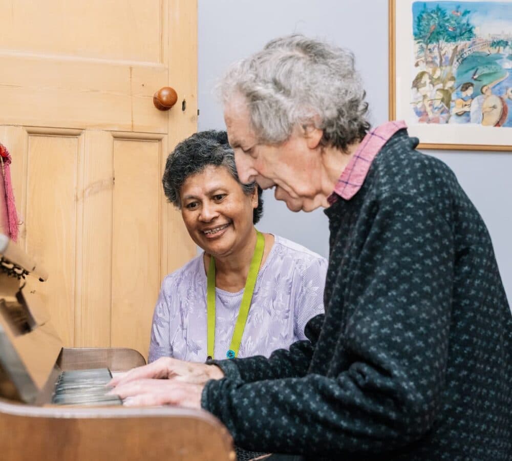 An older male adult with grey hair playing the piano with his younger female carer watching him