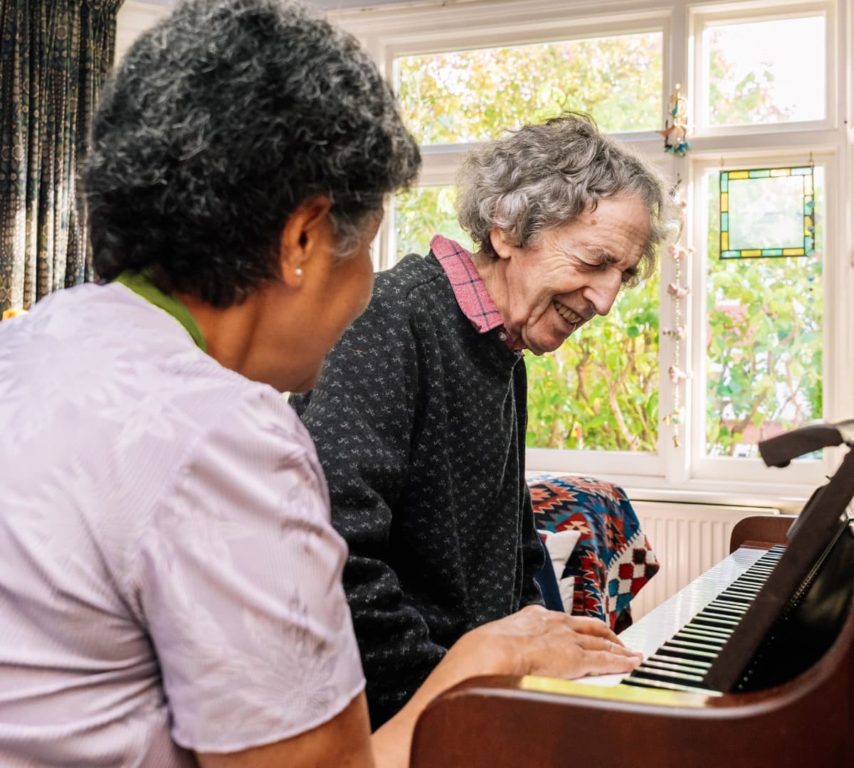An older male adult with short grey hair playing the piano happy and laughing with his female carer with black hair sitting beside him inside the house