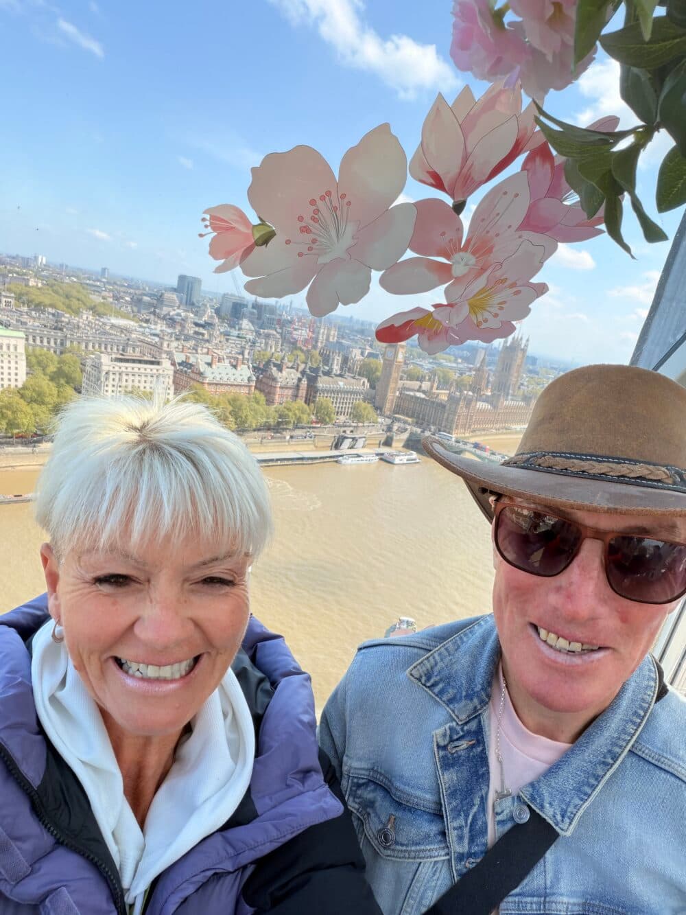 Two smiling people pose on a Ferris wheel with London and Big Ben in the background, flowers above them. - Home Instead