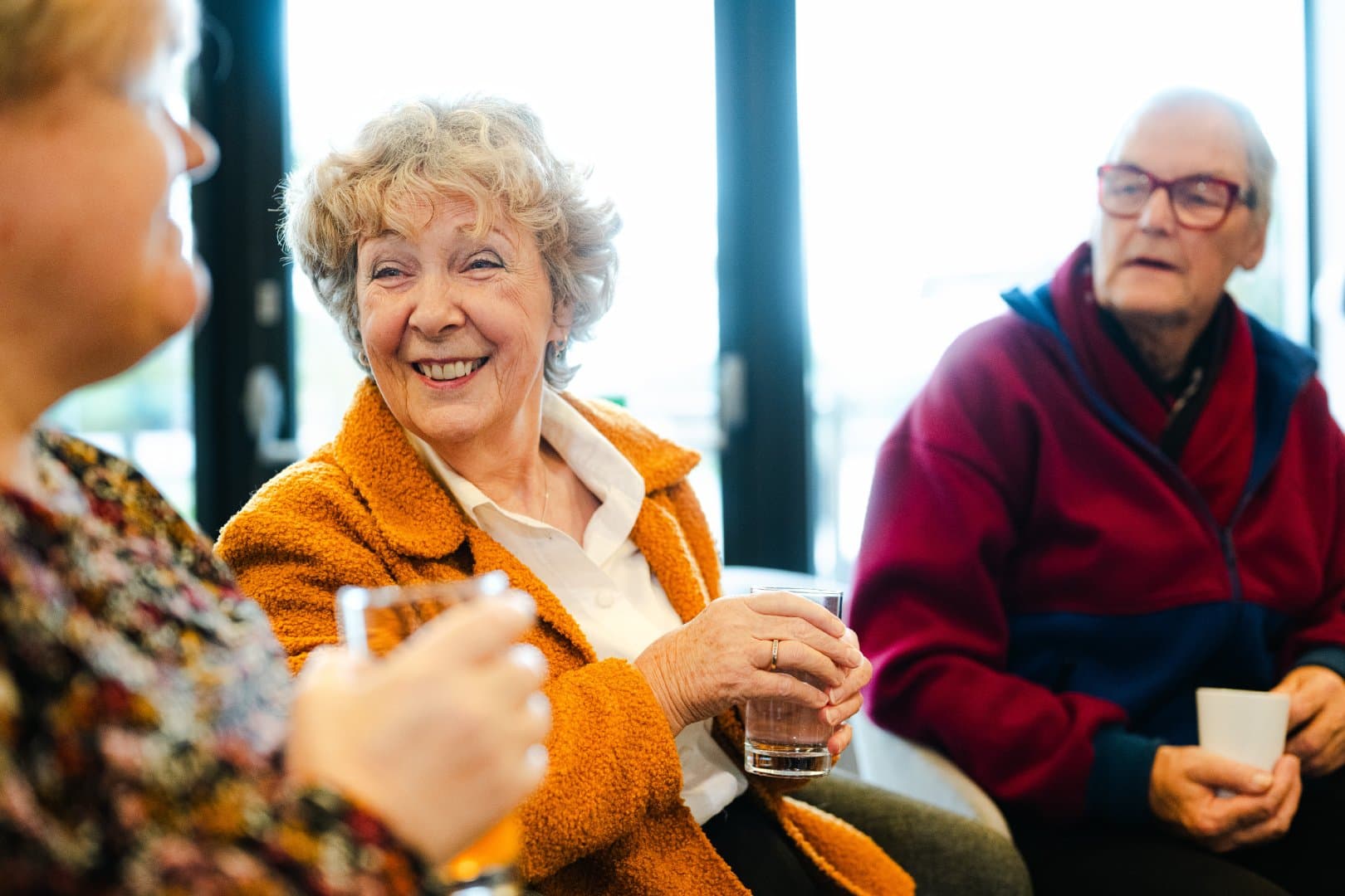 Two older adults smile and chat, holding drinks, seated indoors with another person in the foreground. - Home Instead