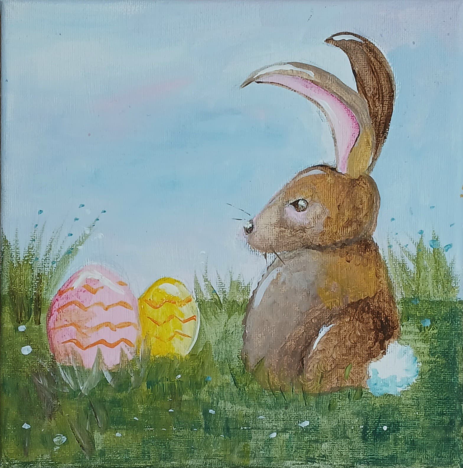 A brown rabbit sits in grass next to two decorated Easter eggs under a blue sky. - Home Instead