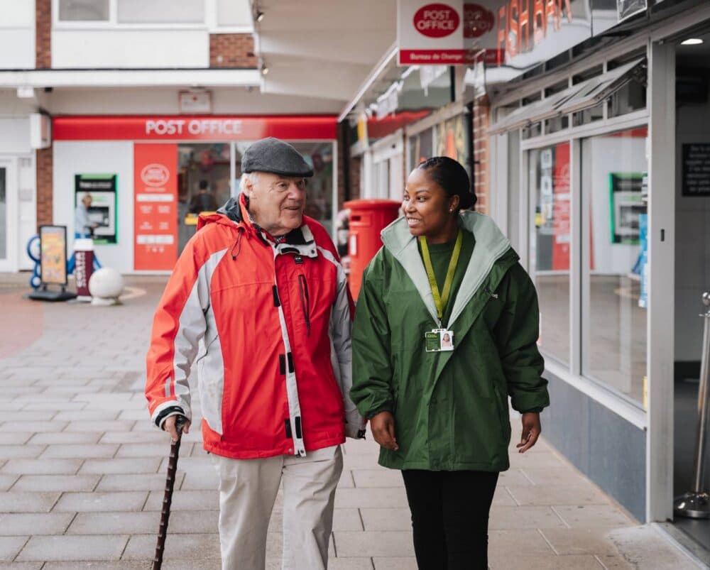 An elderly man with a walking stick walks beside a smiling carer outside a post office. - Home Instead