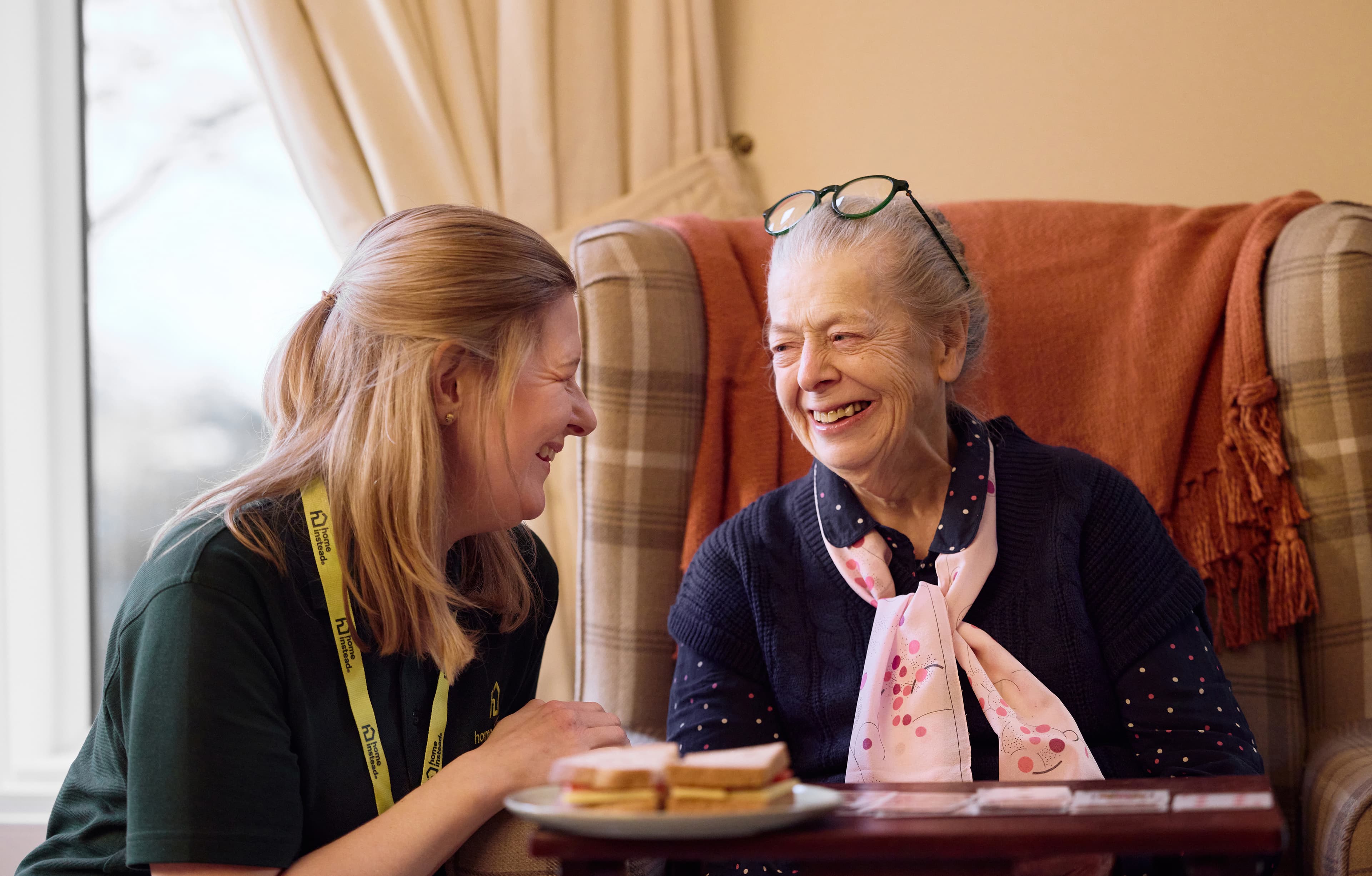 A carer and an elderly woman sit and laugh together, with sandwiches on a table in the foreground. - Home Instead