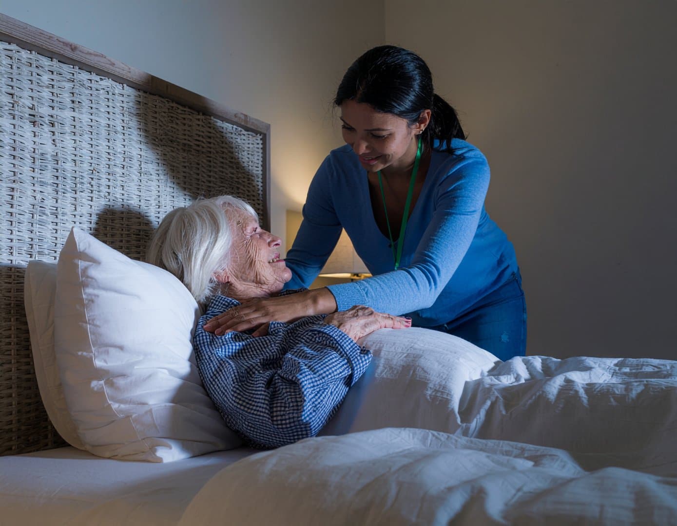 A carer smiles while comforting an elderly woman lying in bed in a softly lit room. - Home Instead