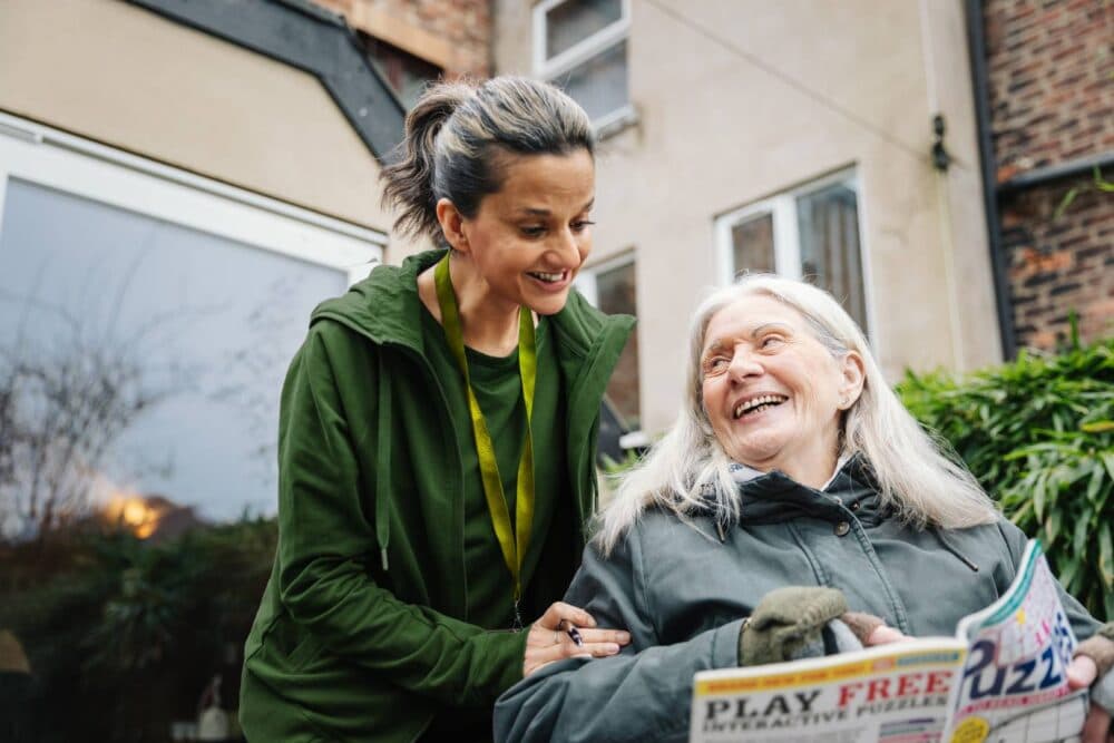 A carer smiles at an elderly woman in a wheelchair holding a puzzle magazine outdoors. - Home Instead